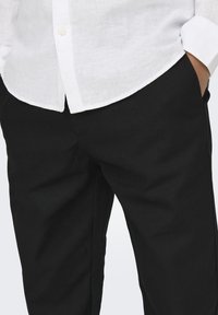 Persona vestita con pantaloni neri e una camicia bianca con bottoni, con una mano in tasca, mostrata dalla vita a metà coscia.