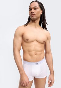 Άνδρας χωρίς πουκάμισο με μακριά κοτσίδα dreadlocks, φορώντας λευκά εσώρουχα Calvin Klein boxer, στέκεται μπροστά σε απλό ανοιχτό γκρι φόντο.