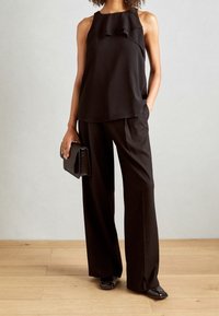Schwarzes ärmelloses Oberteil mit Rüschendetails, kombiniert mit lockeren schwarzen Hosen und einer strukturierten schwarzen Clutch. Auf einem Holzboden arrangiert.