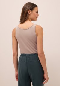 Žena s dlouhými hnědými vlasy oblečená v bezrukávovém světlém mauve topu a tmavě zelených kalhotách s vysokým pasem, stojící na jednobarevném pozadí.