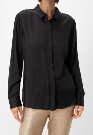 Femme portant une chemise noire à manches longues boutonnée, associée à un pantalon paillettes doré clair brillant sur un fond uni.