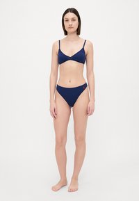 Tums zila bikini komplekts ar trīsstūrveida krūšturi un augsto vidukli, ar smalku viļņotu malu un mazu sarkanu logo detaļu.