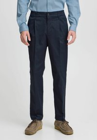 Navy blue pants with pleated front and elastic waistband, made of smooth fabric, paired with light blue long-sleeve shirt and tan loafers.
