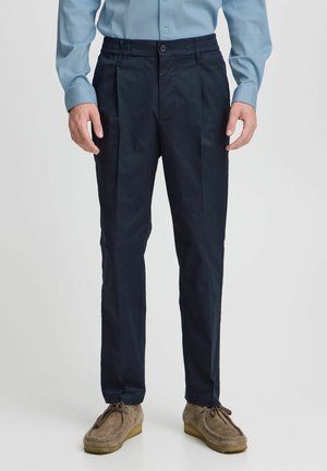 Navy blue pants with pleated front and elastic waistband, made of smooth fabric, paired with light blue long-sleeve shirt and tan loafers.