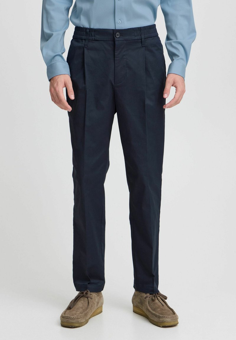 Navy blue pants with pleated front and elastic waistband, made of smooth fabric, paired with light blue long-sleeve shirt and tan loafers.