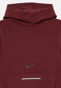 Bordo Nike kapuce ar priekšējo kabatu, atstarojošs pelēks Swoosh logo virs kabatas un atstarojoša horizontāla svītra zem tās.