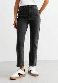 Jeans de mezclilla negra de tiro alto y pierna recta con un sutil desvanecimiento, diseño de cinco bolsillos y cierre de botón, combinados con zapatillas blancas.