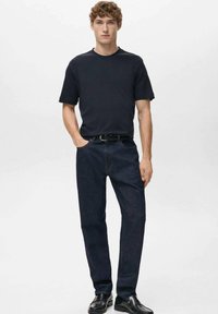 Mlad moški stoji oblečen v črno majico, temno modre kavbojke, črn pas in črne čevlje pred enobarvno svetlo ozadje.