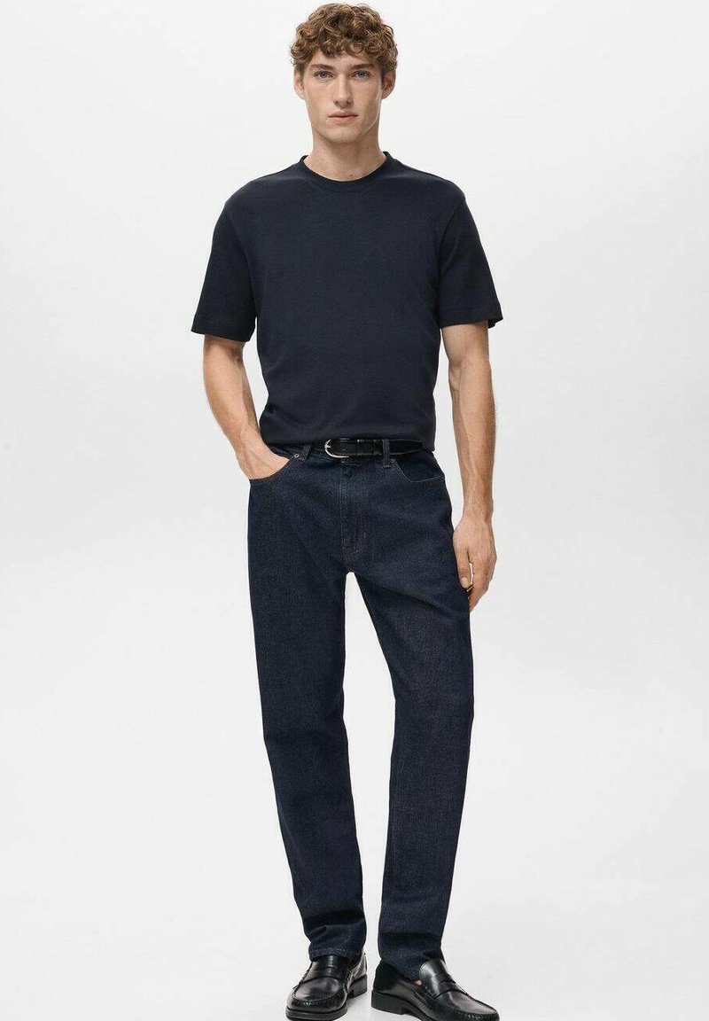 Giovane uomo in piedi che indossa una maglietta nera, jeans blu scuro, una cintura nera e scarpe nere su uno sfondo chiaro.