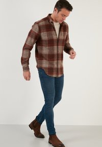 Koszula flanelowa w czerwono-beżowy wzór w kratę, z zapięciem pod szyją, długimi rękawami i zaokrąglonym dołem, zestawiona z wąskimi, ciemnoniebieskimi dżinsami i brązowymi butami.