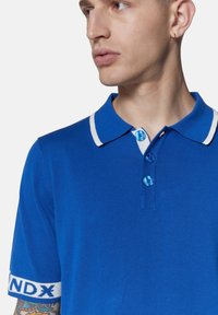 Polo blu con chiusura a due bottoni, collo e polsini bianchi a contrasto, presenta una fascia con logo sul polsino, texture a maglia liscia.