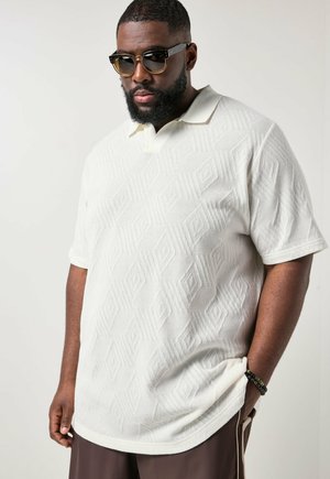 Mann mit Bart und Sonnenbrille, der ein weißes, strukturiertes kurzärmeliges Poloshirt und braune Hose trägt, posiert mit einer Hand in der Tasche.