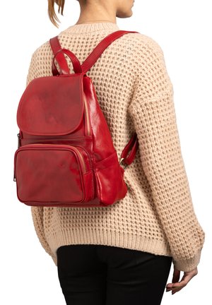 Zaino in pelle rossa con finitura liscia, dotato di un flap superiore, tasche con zip e spallacci regolabili. Indossato su un maglione di lana color crema.