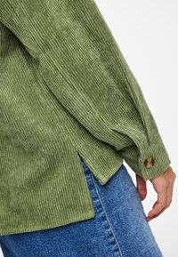 Green corduroy shirt featuring vertical stripes, long sleeves, buttoned cuffs, and side slits. Paired with blue denim.