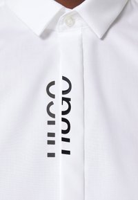 Witte shirt met een klassieke kraag, knoopsluiting en verticale zwarte logo-detail aan de voorkant. Glad materiaal met een frisse textuur.