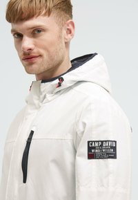 Giacca impermeabile bianca con cappuccio, zip nera, cursori elastici e una patch sulla manica che mostra dettagli del marchio e del design.
