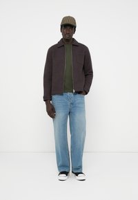 Dark brown zip-up jacket, olive green shirt, light blue wide-leg jeans, and black sneakers with white soles, topped with a plaid cap.