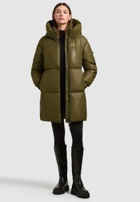 Oliwkowa kurtka puchowa z kapturem, zapinana na zamek, pikowana, z elastycznymi mankietami, zestawiona z czarnymi kozakami za kolano.