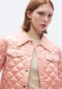 Ροζ μπουφάν με επένδυση καπιτονέ και λείο φινίρισμα, με αντίθετο γιακά, χρυσά μεταλλικά στοιχεία και μπροστινές τσέπες. Φορεμένο πάνω από ριμπ μπεζ τοπ.