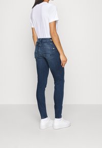 Tummansiniset farkkuleggarit, joissa on korkea vyötärö, takataskut ja valkoinen tikkaus, yhdistettynä yksinkertaiseen valkoiseen t-paitaan ja valkoisiin lenkkareihin.