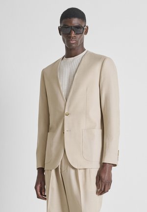 Zwarte man met een oversized beige blazer en broek, crèmekleurige geribbelde trui en grote vierkante zwarte zonnebril, staand tegen een grijze achtergrond.