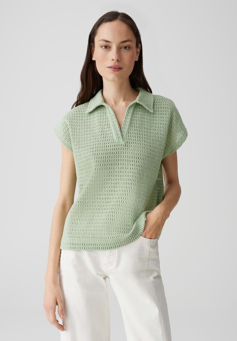 Polo top lavorato a maglia verde chiaro con un motivo testurizzato, maniche corte e colletto, abbinato a pantaloni bianchi.