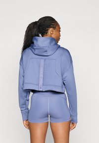 Felpa corta viola con colletto alto e dettagli del logo, abbinata a pantaloni corti sportivi coordinati con una texture liscia e elastica.