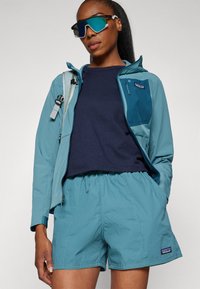 Femeie purtând o geacă verde-albăstruie și pantaloni scurți asortați, cămașă bleumarin, ochelari de soare supradimensionați cu lentile reflectorizante și având o curea de rucsac pe un umăr.