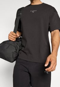 Camiseta de algodón negra con mangas cortas y cuello redondo; logo blanco en el pecho. Sosteniendo una bolsa de gimnasio negra con cremallera y correa ajustable.