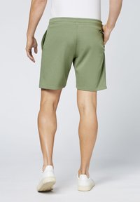 Olivgrüne Sportshorts mit weicher Textur, elastischem Bund und Taschen an den Seiten, kombiniert mit weißen Turnschuhen.