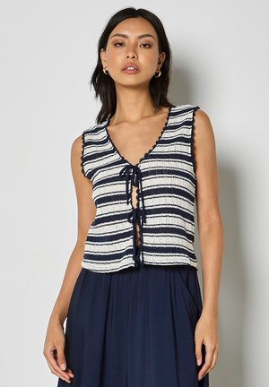 Kobieta w granatowo-białym, bezrękawnym, dzianinowym topie w paski z wiązaniami z przodu i granatową spódnicą z wysokim stanem, stojąca na szarym tle.