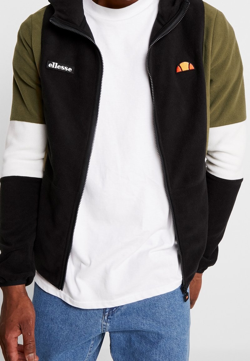 Giacca zip-up in pile nera con maniche verde oliva e bianche. Presenta un logo ricamato e un emblema del sole colorato sul petto.