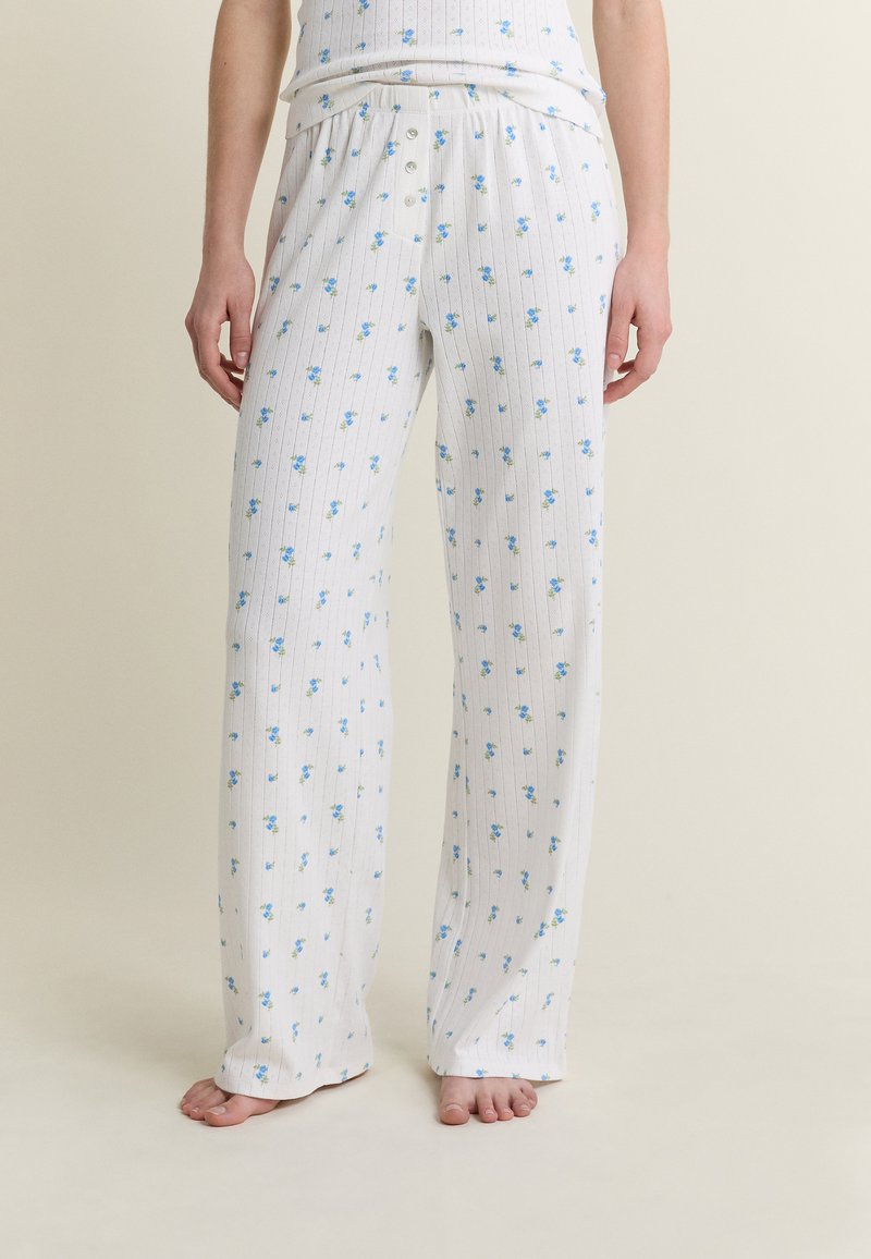 Kobieta w białych spodniach od piżamy z niebieskim kwiatowym wzorem i pionowymi paskami, stojąca boso na jednolitym tle.