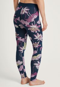 Persona che indossa leggings blu navy con grandi motivi floreali rosa e verde chiaro, standing a piedi nudi su uno sfondo chiaro.