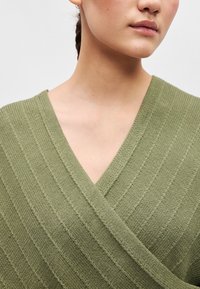 Ελαιόχρωμο πλεκτό περιτύλιγμα με V-λαιμόκοψη, που διαθέτει υφασμάτινες ρίγες και απαλή, υφαντή εμφάνιση. Δεν υπάρχουν ορατοί κλεισίματα.