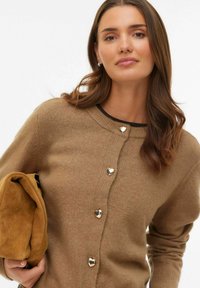 Beige cardigan med hjärtformade silverknappar, ribbad halsringning och slät textur. Modellen håller en brun mockaväska.