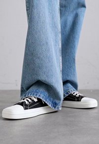 Svarta kanvas sneakers med vita gummisulor och snören, delvis dolda av vida ljusa jeans i denim med en urblekt textur.