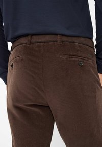 Bruine corduroy broeken met verticale ribbels, met zijzakken en een knoopsluiting. Een donkerblauw shirt is gedeeltelijk zichtbaar boven de taille.