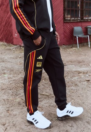Black tracksuit with red and orange stripes, featuring gold accents and logos. Paired with white sneakers featuring black stripes.