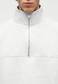 Κοντινό πλάνο ενός ατόμου που φοράει ανοιχτό γκρι πουλόβερ με φερμουάρ μέχρι το στήθος, με το φερμουάρ να είναι μερικώς κλειστό στον γιακά.