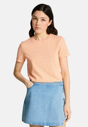 Giovane donna con capelli scuri che indossa una maglietta pesca a maniche corte e una gonna di denim azzurro chiaro, in piedi con una mano in tasca.