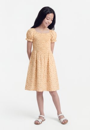 Ragazza giovane con capelli lunghi e scuri, indossa un vestito giallo floreale fino al ginocchio e sandali bianchi, in piedi davanti a uno sfondo bianco.