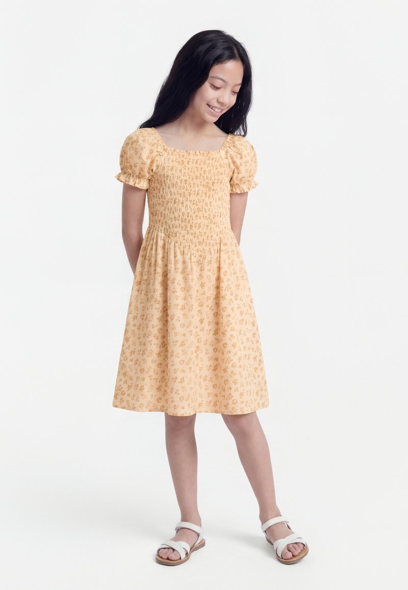 Νεαρή κοπέλα με μακριά σκουρόχρωμα μαλλιά, φορώντας κίτρινο φόρεμα μέχρι τα γόνατα με λουλούδια και άσπρα σανδάλια, στέκεται μπροστά σε λευκό φόντο.