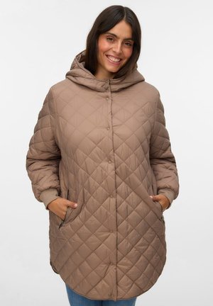 Femme souriante portant un manteau matelassé marron à capuche avec des boutons-pression et les mains dans les poches zippées.