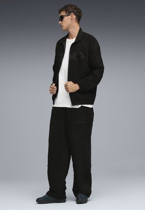 Zwarte suède jas met een hoge kraag, wit T-shirt en loszittende zwarte broek. Accessoires zijn onder andere een zonnebril en schoenen met blauwe accenten.