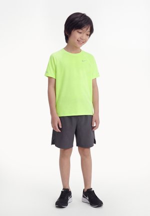 Garçon en chemise à manches courtes vert citron, short gris foncé et chaussures de sport noires, debout devant un fond blanc, souriant et regardant vers le bas.