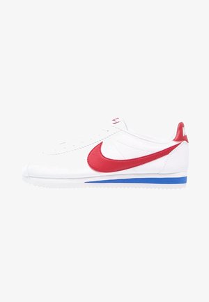Sneaker bianca Nike con logo swoosh rosso, linguetta rossa sul tallone e striscia blu sulla suola bianca, vista laterale, senza sfondo.