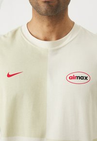 Heledbež t-särk, millel on punane Nike'i silt ja must "aimax" logo punases ovaalis, kahetoonilise disainiga.