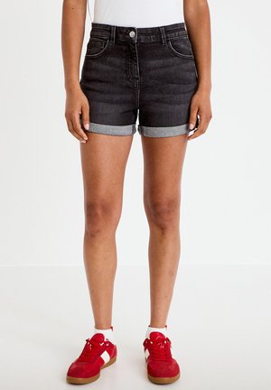 Person iført sorte, op rullede denimshorts, hvide sokker og røde sneakers, stående mod en ensfarvet hvid baggrund.