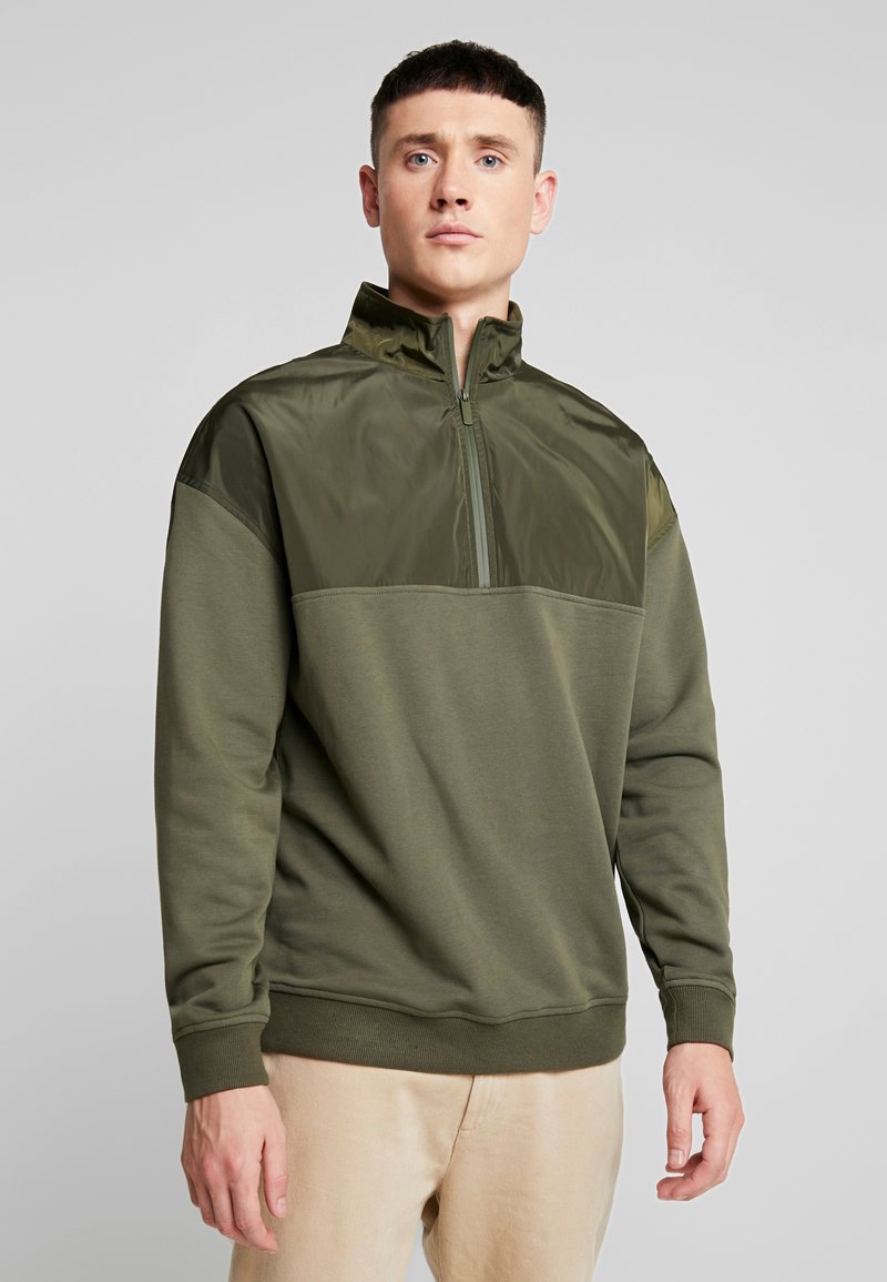 Pullover verde oliva con mezza zip, caratterizzato da un pannello superiore testurizzato e lucido e una sezione inferiore morbida e opaca. Polsini e orlo a coste.